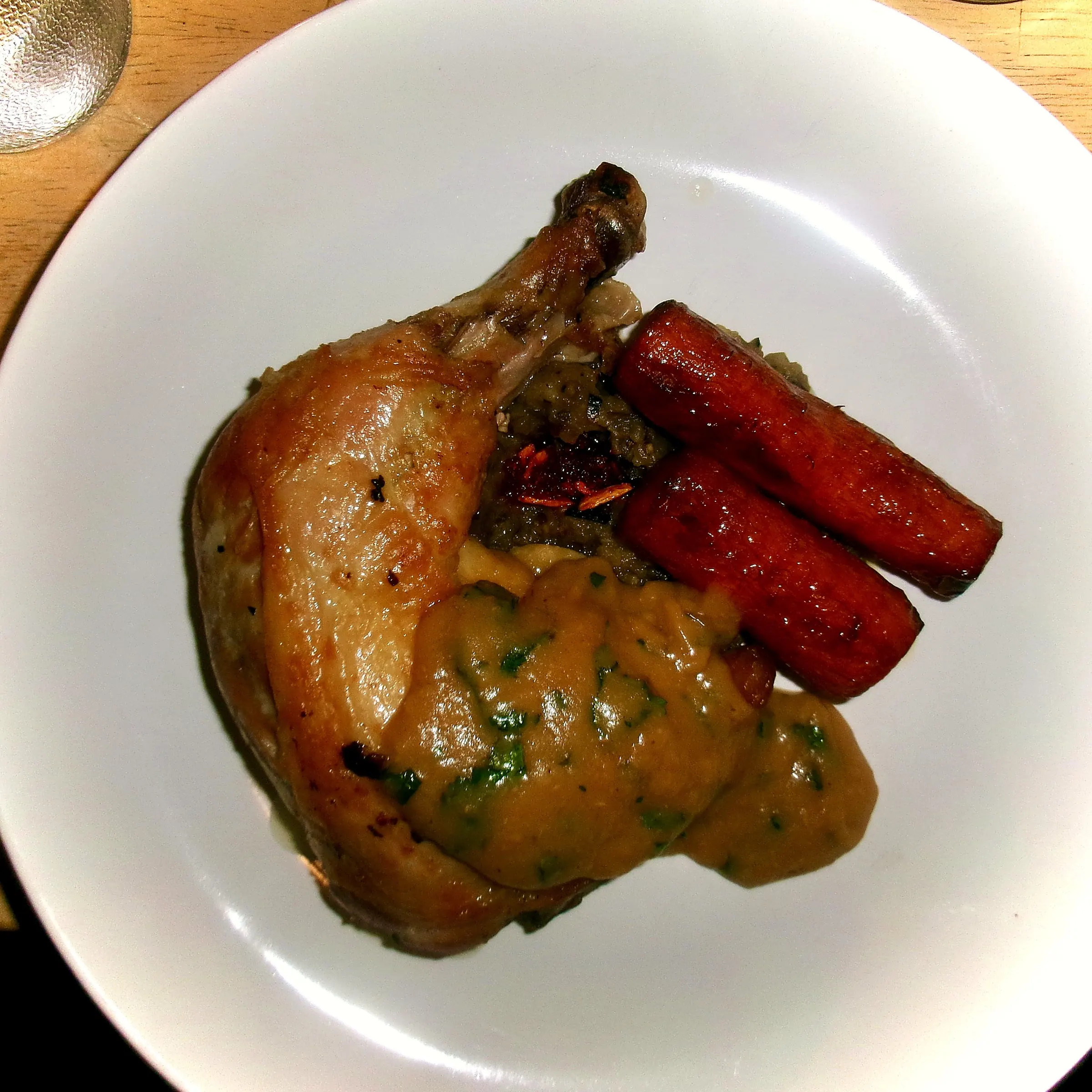 Roast chicken leg under a parsley-flecked pan gravy, with two whole roasted carrots and a dark stuffing, in a wide white bowl.