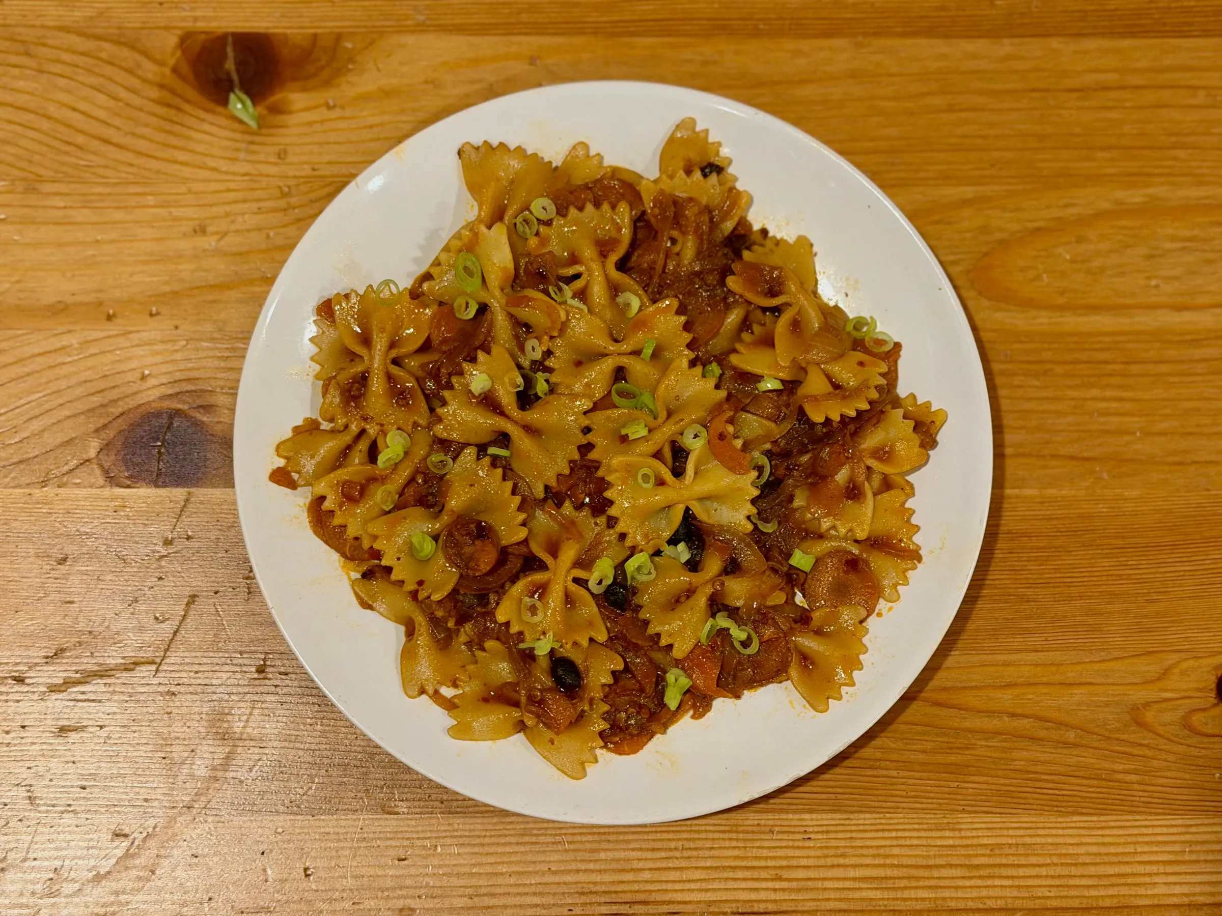 Bowtie pasta in a dark, glossy sauce with sliced sausage, scattered with scallions, on a white plate on a wooden table.