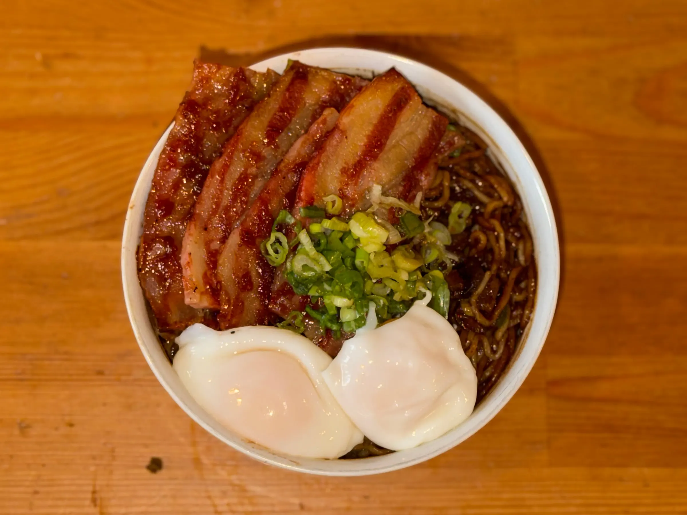 Bowl of dark, soy-glazed noodles topped with slices of grilled pork belly, a heap of scallions, and two poached eggs.