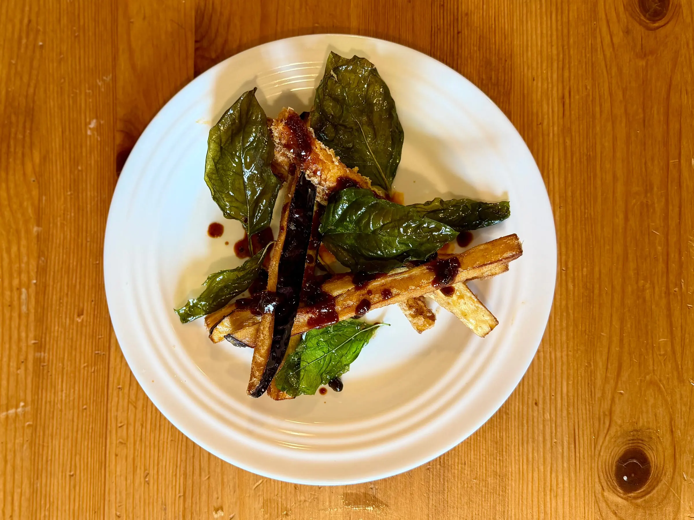 Fried eggplant batons with crisp fried basil leaves and drops of a dark glaze on a ridged white plate.